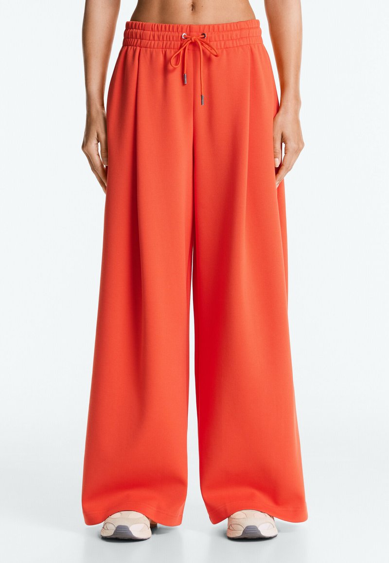 Pantalon orange à jambes larges avec taille élastique et cordon de serrage, porté avec des baskets beiges par une personne debout.