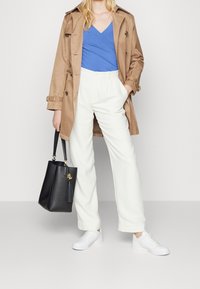 Abrigo trench beige sobre una camiseta azul de cuello en V, pantalones anchos blancos, zapatillas blancas y un bolso negro con un detalle dorado.