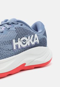 Pantof de sport albastru cu partea superioară din plasă texturată, logo-ul alb proeminent "HOKA", talpa albă căptușită cu accente roșii și detalii de design stratificate.