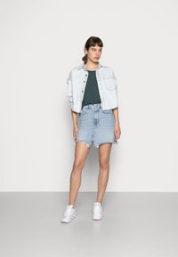 Hellblaues Denim-Jacket, grünes Oberteil, distressed hellblauer Denim-Rock mit fransigem Saum, weiße Sneakers mit pastellfarbenen Akzenten.