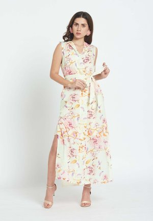 Ženska si prilagaja pas na brezrokavnem maxi obleki s cvetličnim vzorcem in bočnim razporkom, obuta v bež pete in z rožnatimi opaznimi uhani.