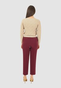 Donna che indossa un pullover beige testurizzato, pantaloni bordeaux e sandali dorati con tacchi alti, in piedi con la schiena rivolta verso uno sfondo neutro.