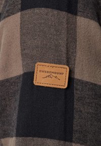 Tissu à carreaux marron et noir avec un patch en cuir cousu portant l'inscription « Swedemount » et un logo de montagne au centre.