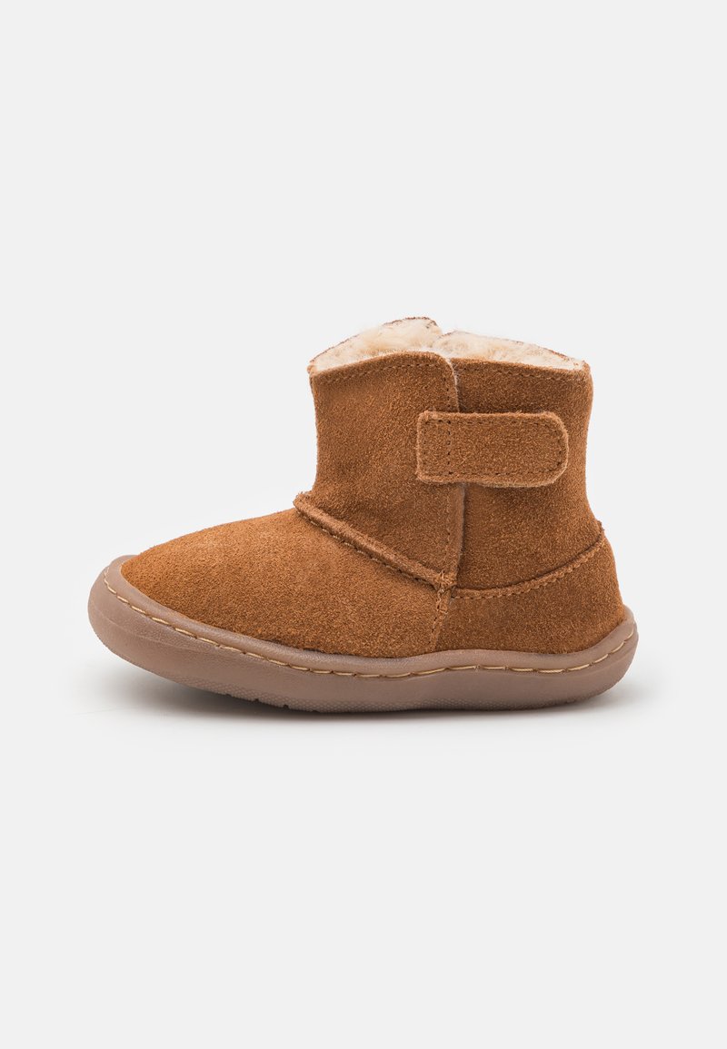 Botín de ante marrón con un suave forro de piel de pelo, cinta de Velcro, puntera redondeada y suela de goma flexible. Detalle de costura ligera a lo largo del borde.