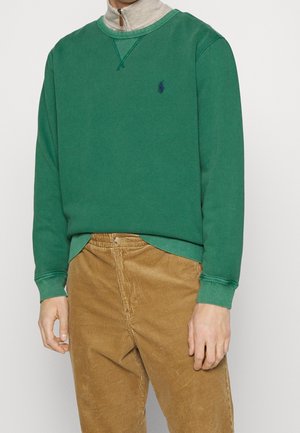 Mand iført grøn langærmet sweatshirt over beige lynlåstop og lysebrune kordbukser, stående mod en ensfarvet baggrund.