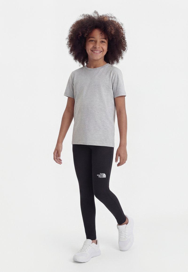 Enfant souriant aux cheveux bouclés portant un t-shirt à manches courtes gris clair, des leggings noirs avec le logo The North Face, et des baskets blanches, marchant vers l'avant.