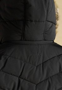 Mustard riidest jope kapuutsiga, mille ülemisel seljal kaela all on broderitud brändinimi "JOULES".