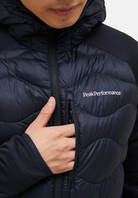 Marineblå luftmadrasjakke med et quiltet mønster, der har en høj krave, lynlås lukning og logo på brystet. Tekstureret stof og tætsiddende manchetter.