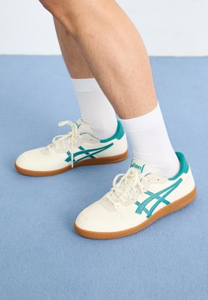 Person wearing white and teal Asics sneakers with gum soles and white crew socks standing on a light blue carpet.