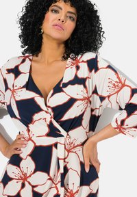 Abito a portafoglio floreale in blu navy e panna, con grandi fiori rossi e bianchi, scollo a V profondo, maniche a tre quarti e dettaglio in vita arricciata.