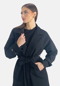 Czarny trenchcoat wykonany z fakturowanej tkaniny, z paskiem w talii, szerokim kołnierzem i zapinanymi mankietami. Minimalistyczny design z funkcjonalnymi kieszeniami.