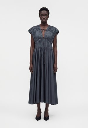 Kobieta stojąca, ubrana w bezrękawnikową, ciemnoszarą sukienkę midi z marszczonym materiałem i wiązanym dekoltem z wycięciem w kształcie klucza, zestawioną z czarnymi, spiczastymi szpilkami.