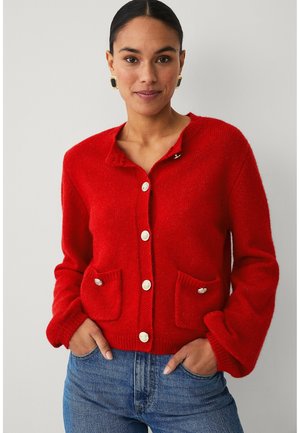 Frau trägt roten, geknöpften Cardigan mit zwei Fronttaschen und blaue Jeans, steht vor schlichtem hellem Hintergrund.