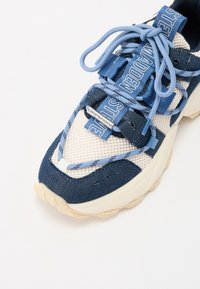 Objemné tenisky s pletenou svrškem v krémové a námořnické modré barvě, s texturovanými prvky a modrými šnurbatky. Mají pohodlnou béžovou podrážku.