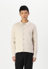 Light beige button-up knit shirt with a collar and a relaxed fit. Features a textured finish and five darker buttons along the front.