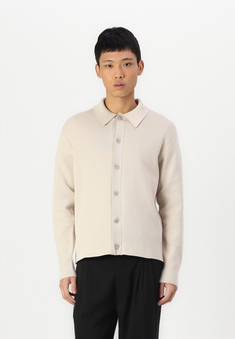 Light beige button-up knit shirt with a collar and a relaxed fit. Features a textured finish and five darker buttons along the front.