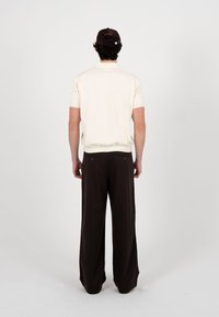 Homme portant une chemise crème à manches courtes, un pantalon large foncé et une casquette de baseball foncée, debout de dos sur un fond uni.