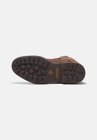 Zapato de ante marrón con una suela de goma duradera que presenta patrones de tracción multidireccional. La suela incluye un logo y detalles texturizados.