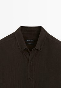 Dunkelbraunes Button-up-Hemd mit strukturiertem Kragen, vertikaler Textur und schwarzen Knöpfen. Etikett sichtbar im Kragen mit Markenname.