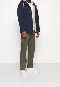 Mörkblå jacka med huva, två framfickor och beige foder; bärs över en vit t-shirt och matchas med olivgröna byxor och vita sneakers.