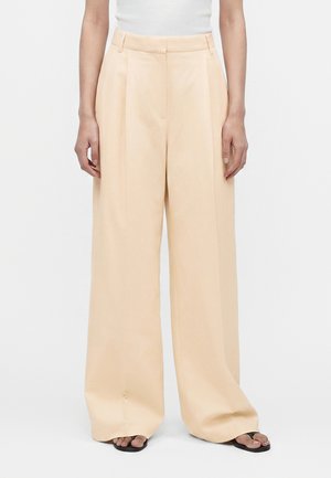 Femme portant un pantalon beige taille haute, jambes larges avec plis et des sandales noires, debout devant un fond blanc uni.