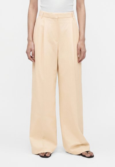 Femme portant un pantalon beige taille haute, jambes larges avec plis et des sandales noires, debout devant un fond blanc uni.