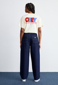 T-shirt em creme suave com texto gráfico vermelho e azul "OBEY ART DEPARTMENT" nas costas; calças de ganga de perna larga de cintura alta em azul escuro; ténis brancos.