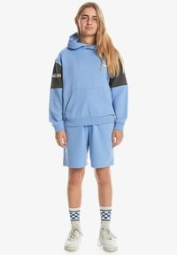 Haut à capuche bleu clair avec des accents noirs, poche kangourou, associé à un short assorti. Des baskets blanches et des chaussettes à motifs complètent le look.