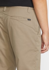 Los pantalones cargo beige cuentan con un bolsillo lateral y una etiqueta negra de "Volcom". La tela tiene una textura suave con sutiles detalles de costura.