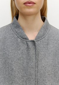 Grijze wolmixjas met een hoge kraag en minimale naaddetails, met een zachte textuur en een knoopsluiting bij de hals.