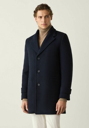 Mann trägt einen marineblauen Wollmantel über einem beigen Rollkragenpullover und weißen Hosen, steht vor einem einfarbigen Hintergrund.