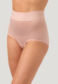 Donna che indossa slip modellanti senza cuciture a vita alta, di un delicato rosa chiaro, su uno sfondo chiaro e uniforme.