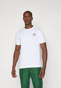 Λευκό βαμβακερό t-shirt με στρογγυλή λαιμόκοψη και κοντά μανίκια, με μαύρη εκτύπωση λογότυπου στο αριστερό στήθος. Φορεμένο με πράσινα παντελόνια.