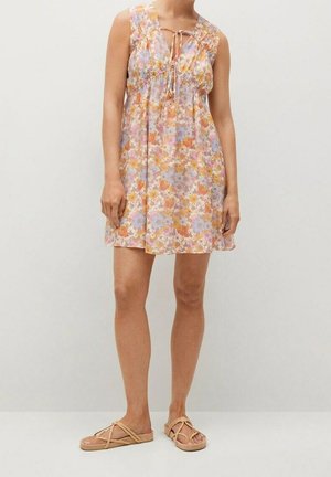 Kvinde iført en ærmeløs kort kjole med blomstermønster og bindedetalje ved udskæringen samt beige flade sandaler med flettede remme.