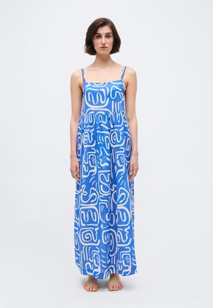 Žena v modrých maxi šatách bez rukávov s bielymi abstraktnými vzormi, stojaca bosá na čisto bielom pozadí.