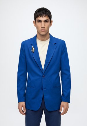 Homme portant un blazer bleu vif avec une broche dorée en forme de main sur une chemise blanche, debout devant un fond clair uni.