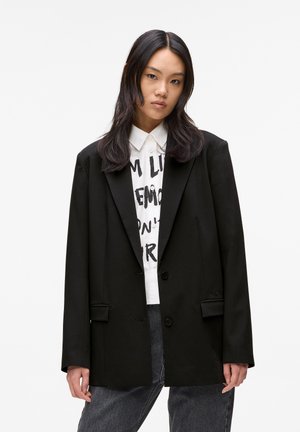 Young woman with long dark hair wears oversized black blazer, white shirt with black text, and dark gray jeans against a plain white background.