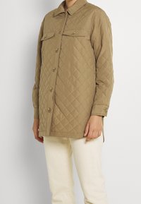 Gequilted tan jas met een kraag, voorknopjes en twee borstzakken. Heeft lange mouwen en een licht ontspannen pasvorm.