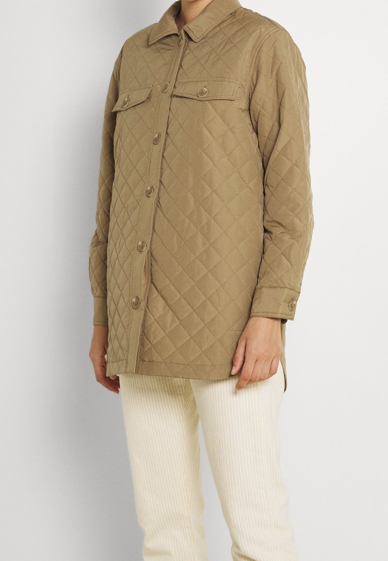 Gequilted tan jas met een kraag, voorknopjes en twee borstzakken. Heeft lange mouwen en een licht ontspannen pasvorm.