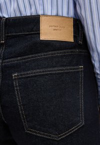 Κοντινό πλάνο από πίσω τσέπη σκούρων μπλε τζιν με ταμπέλα σε ταμπά χρώμα που γράφει "perfect jeans gina tricot", φορεμένα με μπλε και λευκή ριγέ πουκαμίσα.