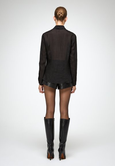 Camicia nera trasparente a maniche lunghe con taglio dritto, indossata sopra pantaloni corti neri lucidi. Il modello indossa stivali neri con tacco alto, con una texture liscia.