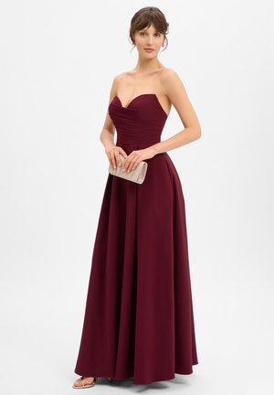 Burgunderfarbener, trägerloser Abendkleid mit tailliertem Oberteil und fließendem Rock. Model hält eine metallische Clutch und trägt Riemchen-Absätze.