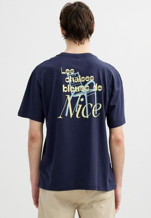 Hombre con camiseta azul marino con texto beige y gráfico de una silla azul que dice "Les chaises bleues de Nice" en la espalda.