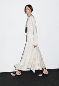 Wijds, wit maxirok met een lichte textuur, gecombineerd met een top met lange mouwen. Zwarte sandalen met bandjes en een hangende ketting maken de look compleet.