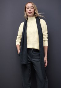 Kremowy sweter z dzianiny o teksturowanym wzorze, sparowany z ciemnoszarym wełnianym szalikiem i dopasowanymi szarymi spodniami. Jednolite kolory, luźny krój.