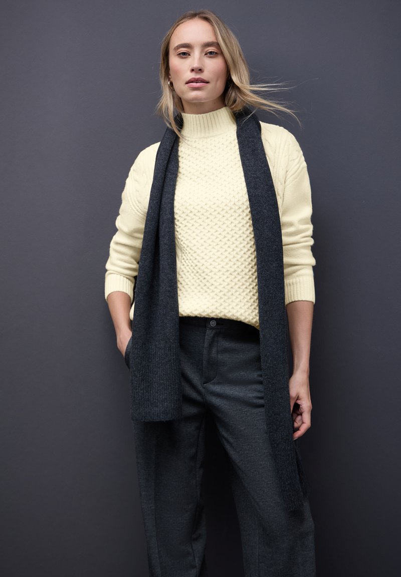 Kremowy sweter z dzianiny o teksturowanym wzorze, sparowany z ciemnoszarym wełnianym szalikiem i dopasowanymi szarymi spodniami. Jednolite kolory, luźny krój.