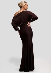 Brun axelbandslös lång klänning i mjukt tyg, med en figurnära överdel och en flödande kjol. Långa ärmar med rynkad detalj.