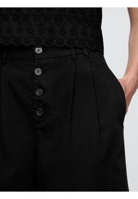 Pantalons taille haute noirs avec des plis, dotés de grands boutons et de poches latérales. Le tissu a une texture lisse avec un léger éclat.