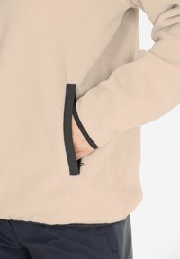 Beige fleecejack met zwart accent, voorzien van een zijzak en elastische boorden. Het materiaal lijkt zacht met een gladde textuur.