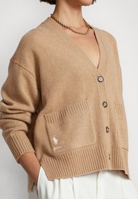 Beige stickad kofta med V-ringning, knappar fram, ribbade muddar och två framfickor; har en liten broderad logotyp på fickan.